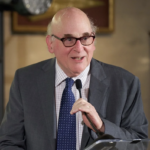 Paul Aisen is wearing a grey suit and holding a microphone at a podium